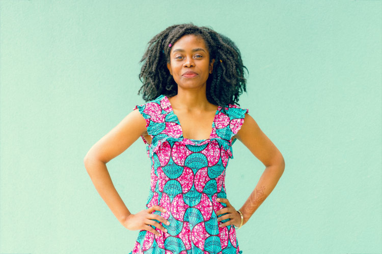 Woman with dark curly hair wears a pink and teal dress with her hands on her hips